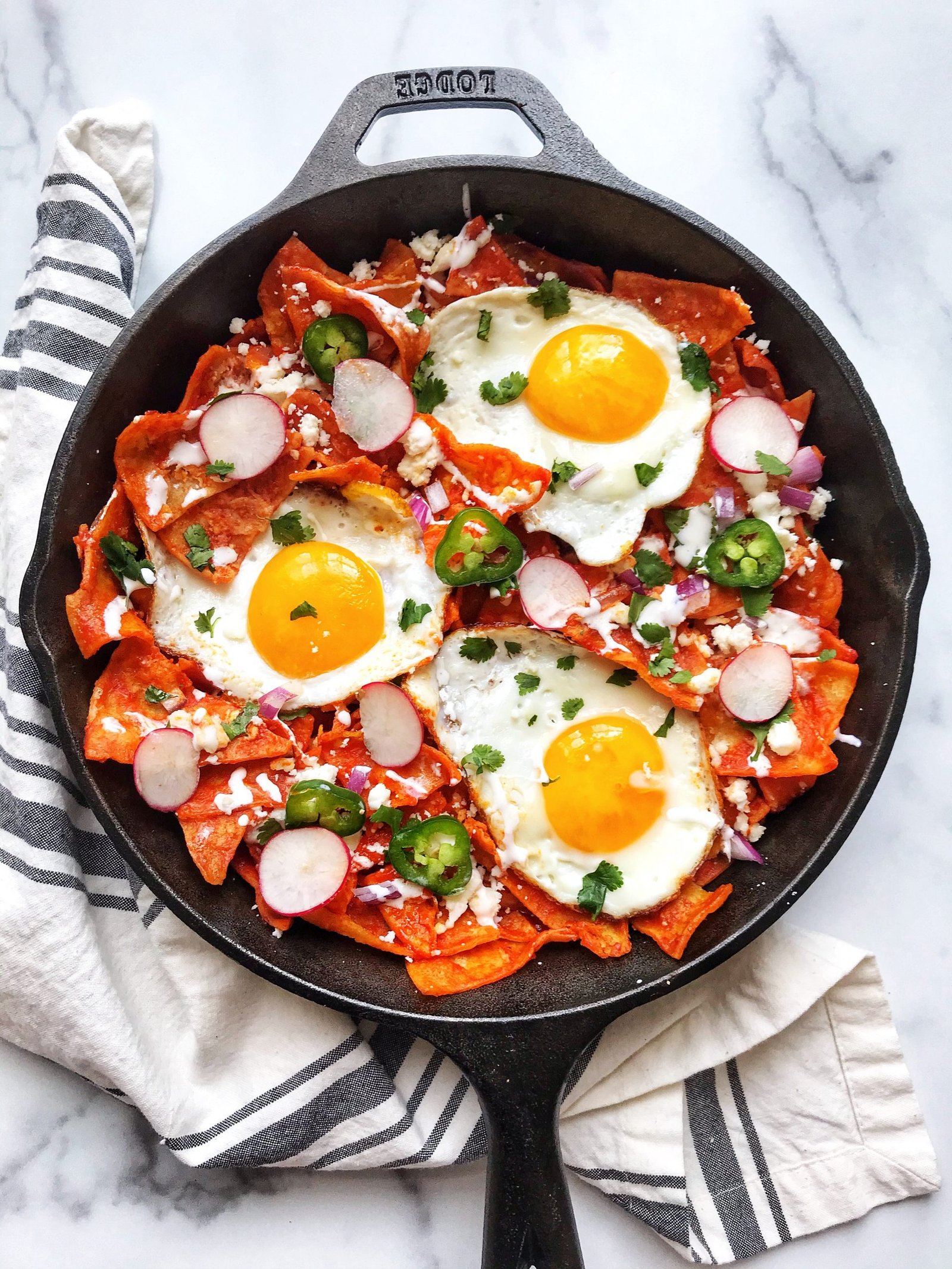 Chilaquiles Rojos (Red Chilaquiles) in cast iron skillet with cheese, sour cream, onions, jalapenos, fried eggs