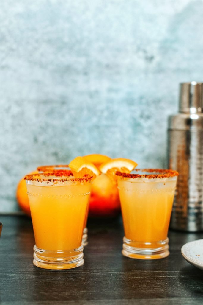 Three orange mango margaritas in small glass with Tajin rim. Shaker in background.
