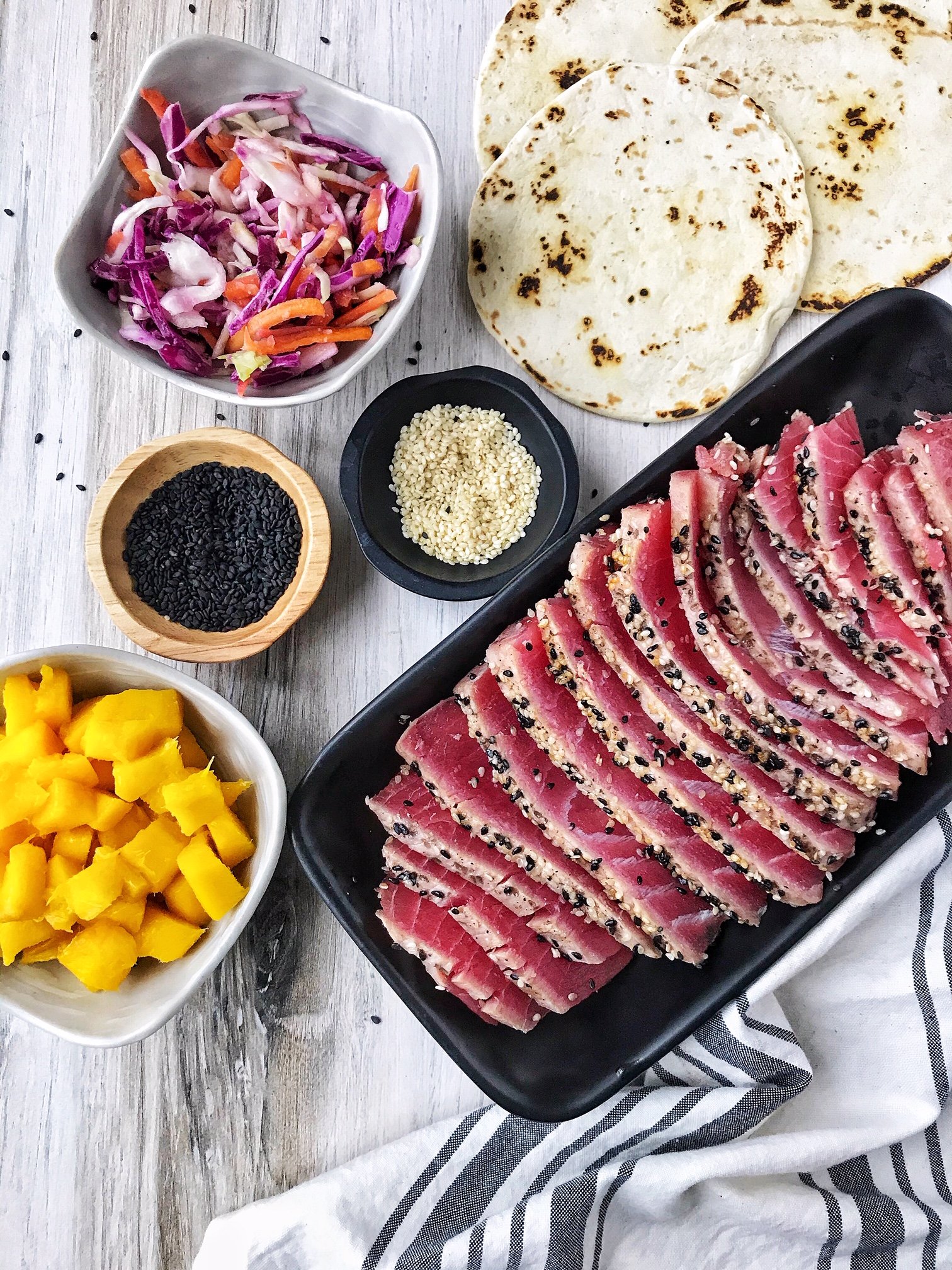 Seared tuna on black plate. Black and white sesame seeds and slaw on the side. Charred flour tortillas.