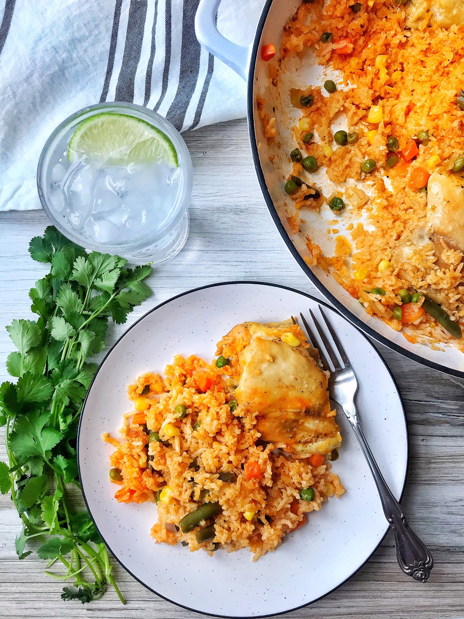 Large pan of chicken and rice. Small plate with chicken thigh and rice with vegetables. Glass of iced water with lime. Cilantro on the side.