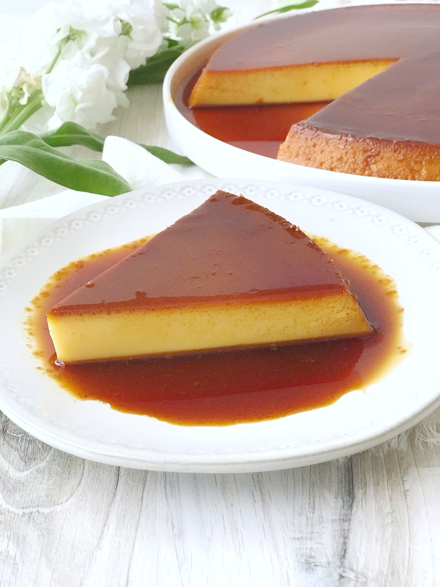 Mexican flan on large white plate with slice missing. Mexican flan slice on small white plate with caramel sauce. White flowers.