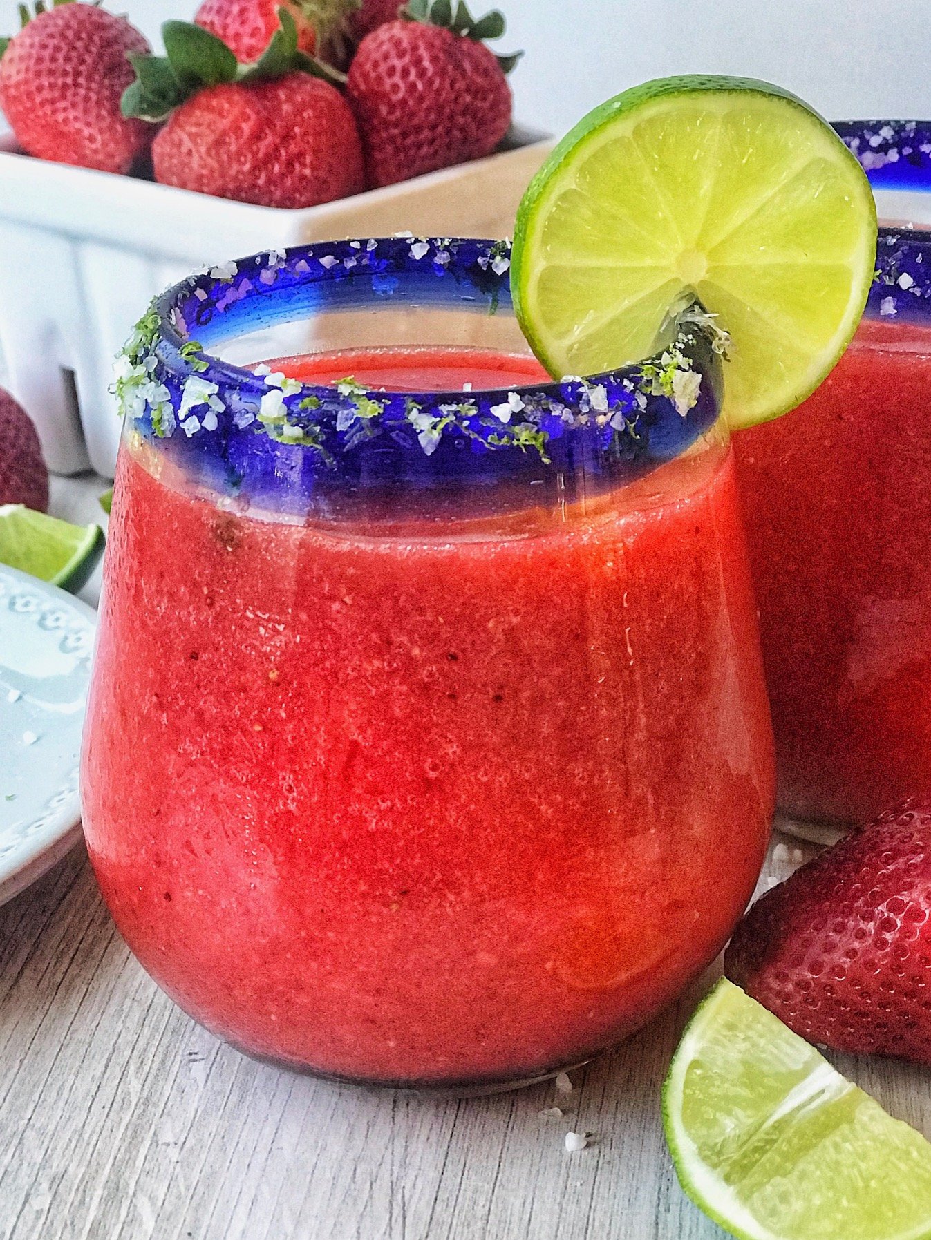 Frozen strawberry margarita in a glass with blue rim. Salt and lime zest on rim with lime slice garnish. Strawberries in the background.