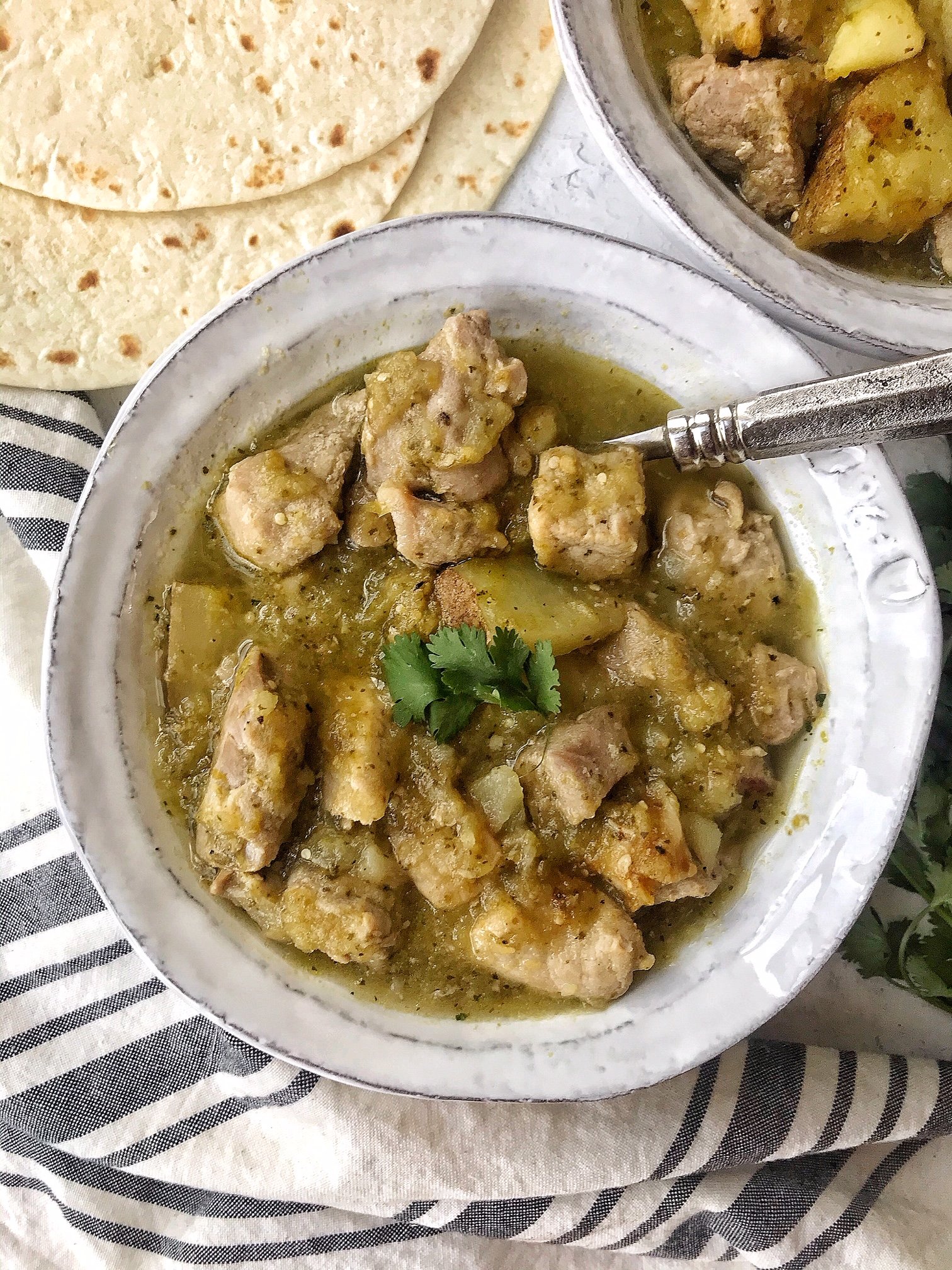 White bowl with pork chile verde and silver spoon. Flour tortillas on the side.
