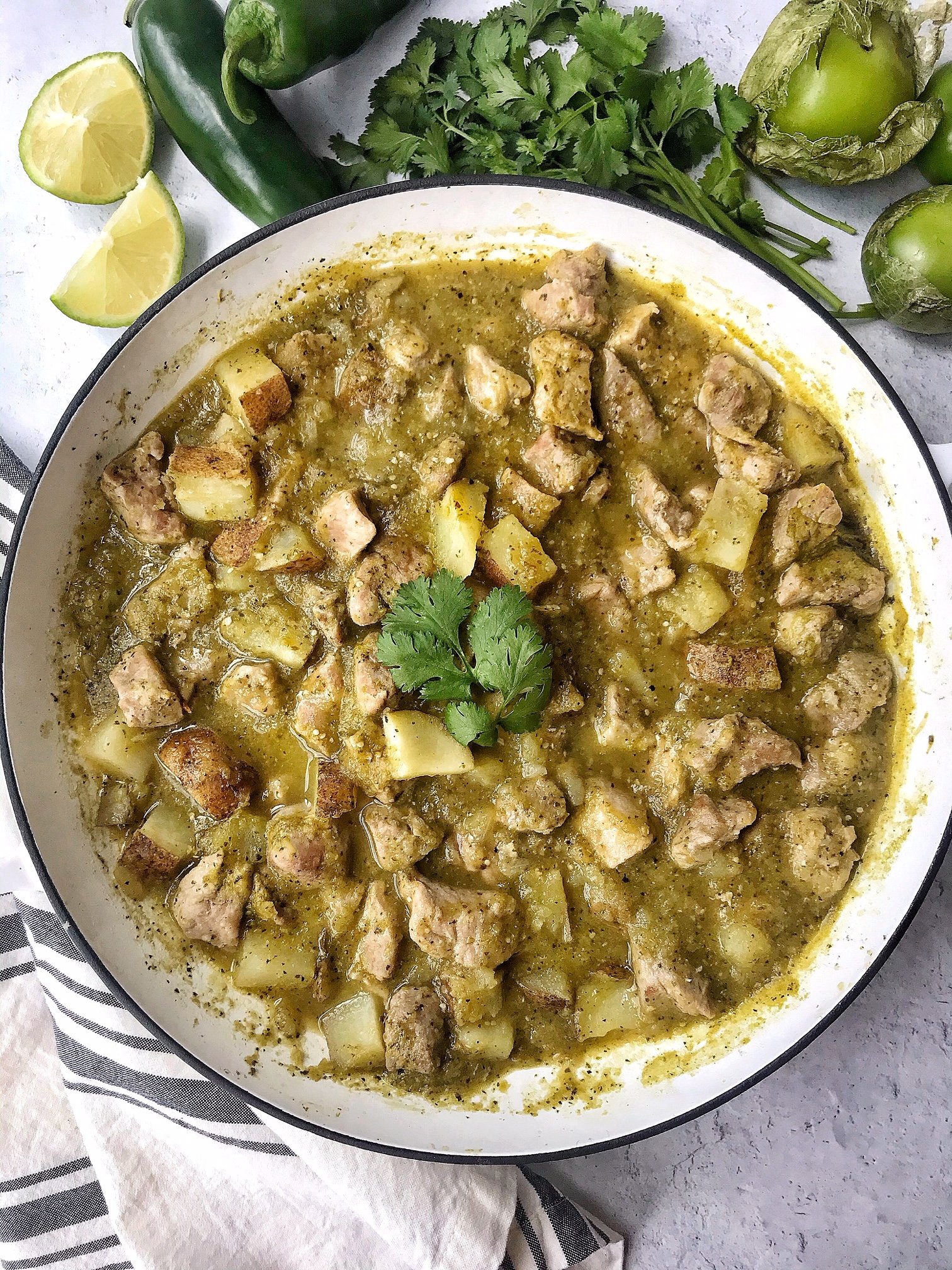 Pork chile verde in a white skillet. Jalapenos, tomatillos, lime, and cilantro as props