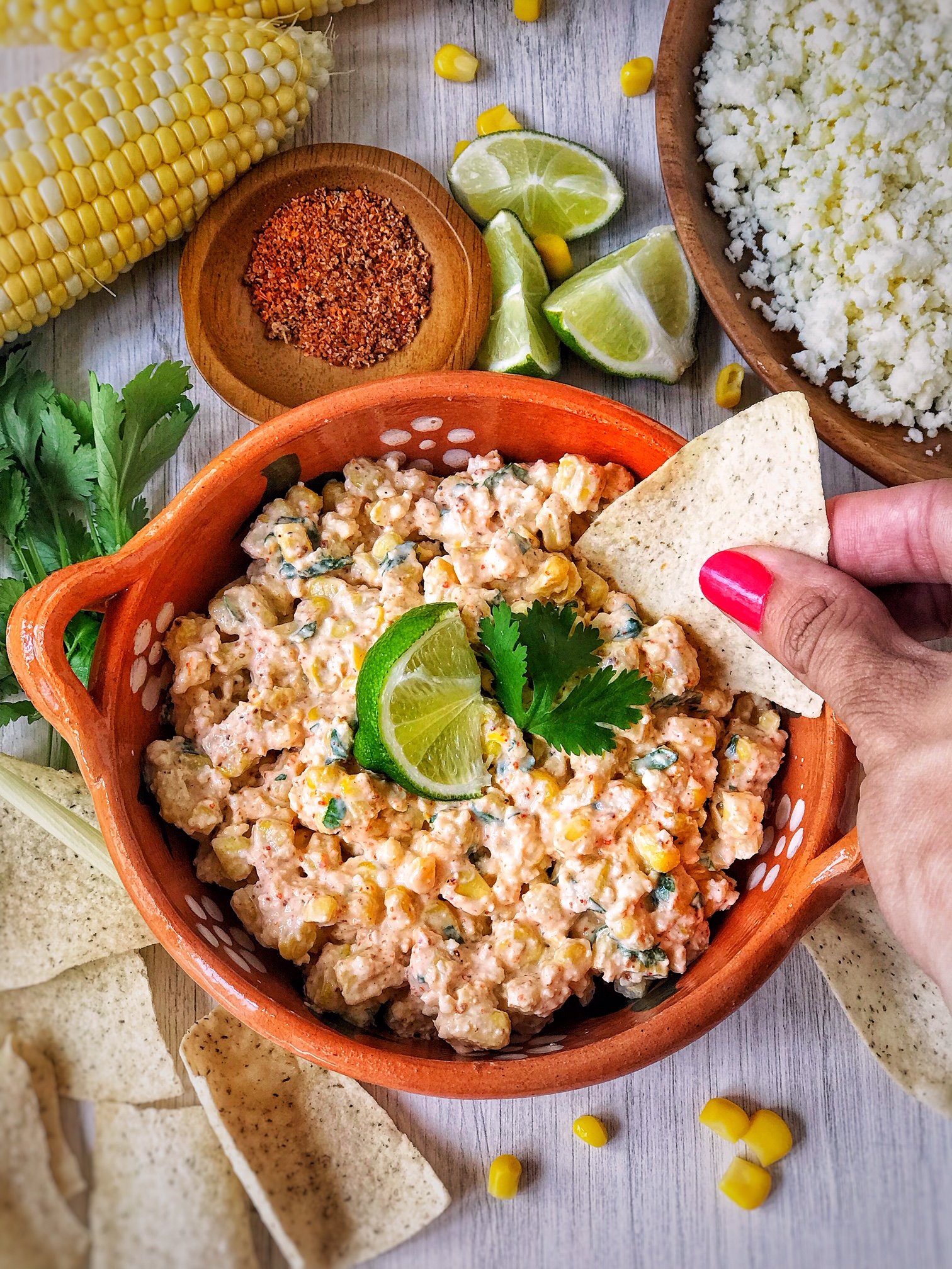Bowl of Mexican street corn dip garnished with lime and cilantro. Hand in picture with tortilla chip in dip. Corn, limes, and crumbled cheese in picture.