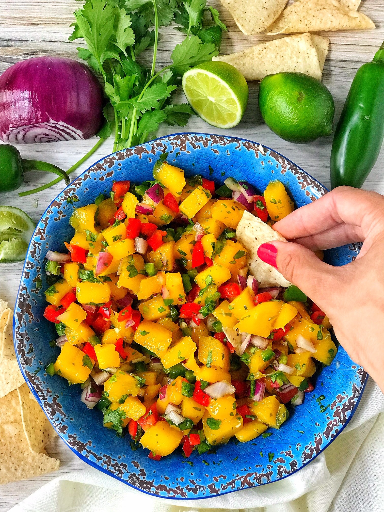 Blue bowl with mango salsa including mango, cilantro, red onion, red bell pepper. Hand dipping tortilla chip into mango salsa. Chips, cilantro, red onion, limes, and jalapeno in picture.