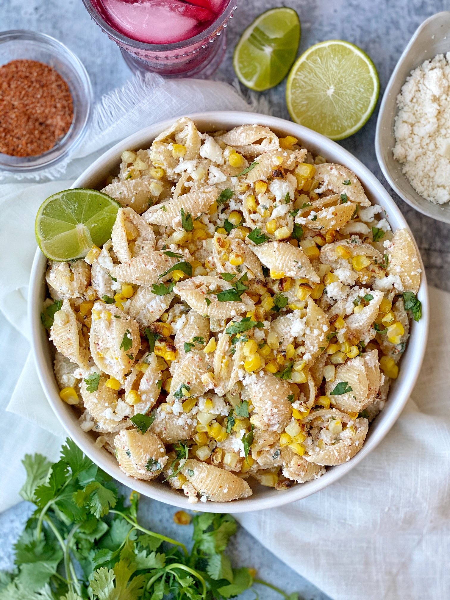 Pasta salad with corn in a white plate with lime garnish. chili powder on side, crumbled cheese on side.