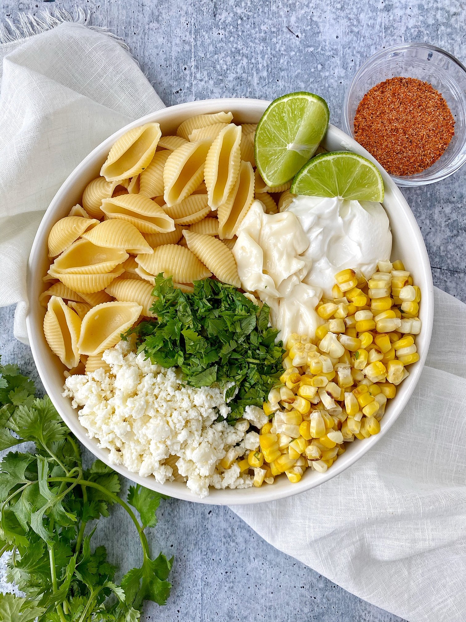 Pasta, sour cream, crumbled cheese, cilantro, and corn in a white bowl. Lime garnish, cilantro garnish, chili powder on the side.