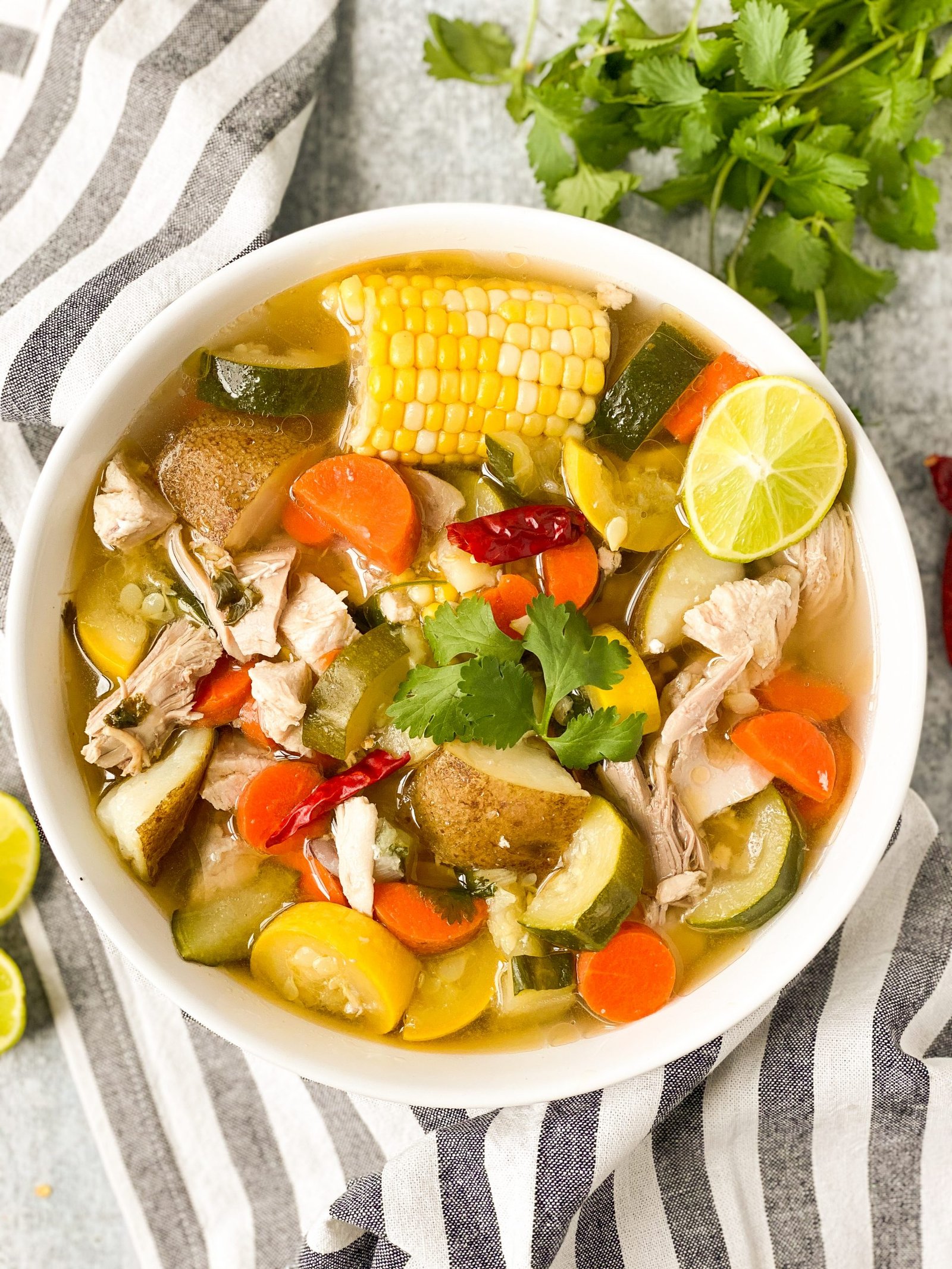 White bowl with caldo de pollo, mexican chicken soup. Includes potatoes, zucchini, carrots, and corn. Striped towel under bowl.