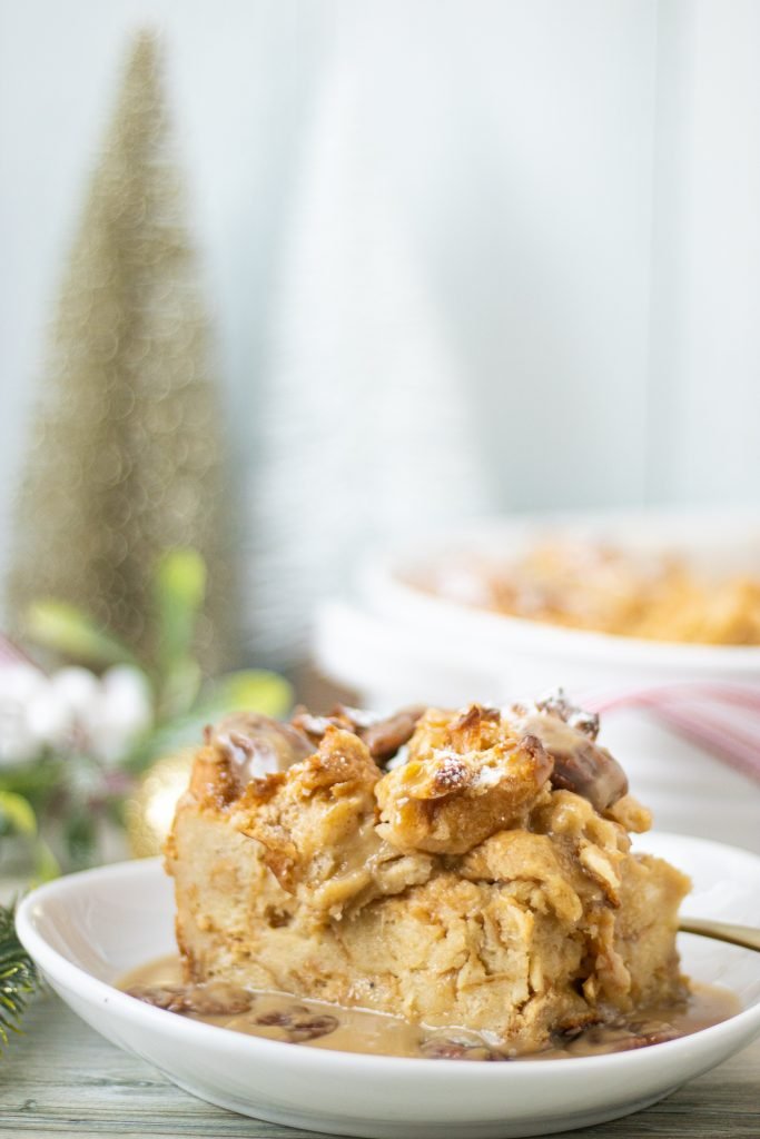 Slice of bread pudding on white bowl with brown pecan sauce. Christmas tree in background