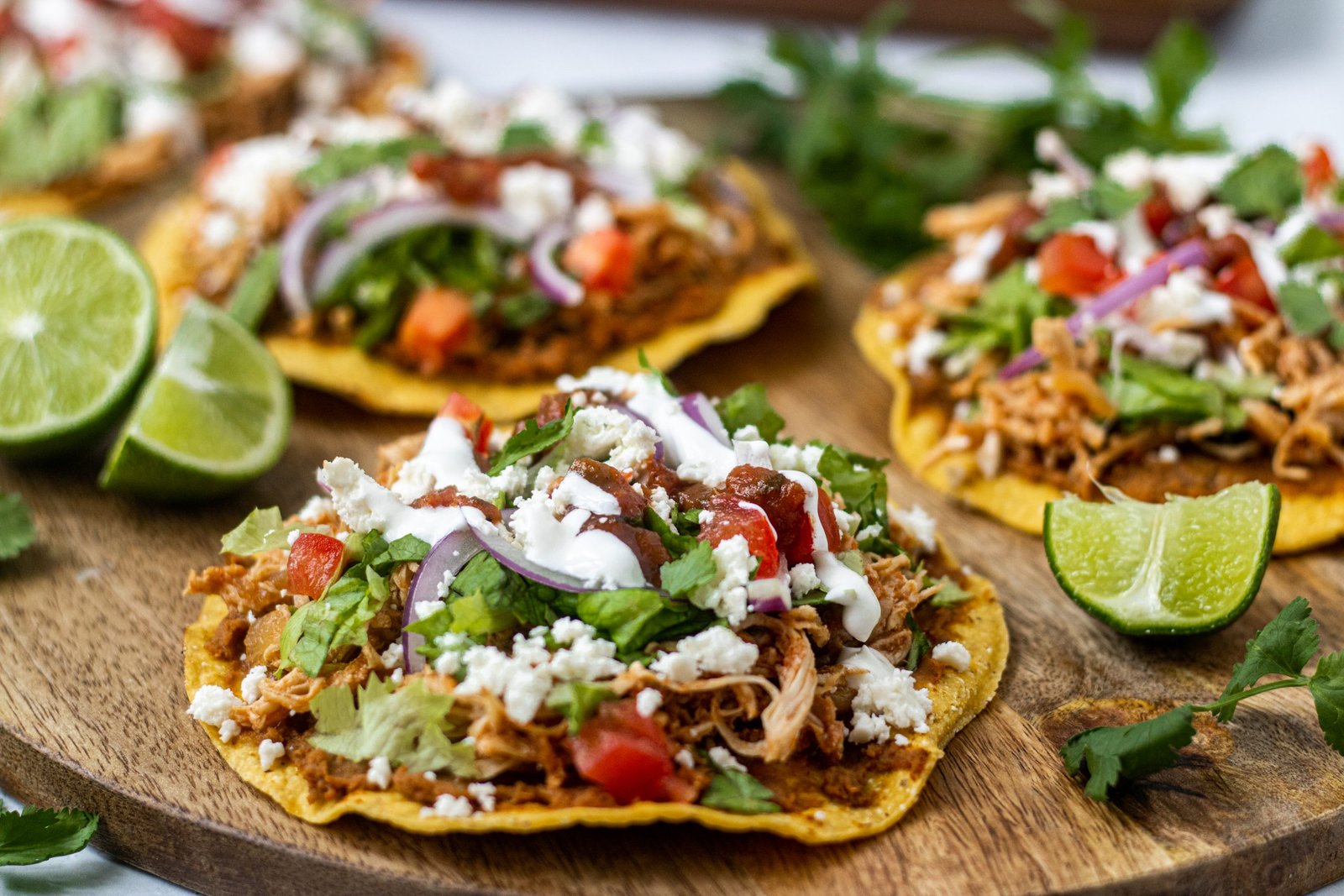 Chicken Tinga Tostadas on wooden tray. Topped with lettuce, onion, tomato, cheese. Lime garnishes