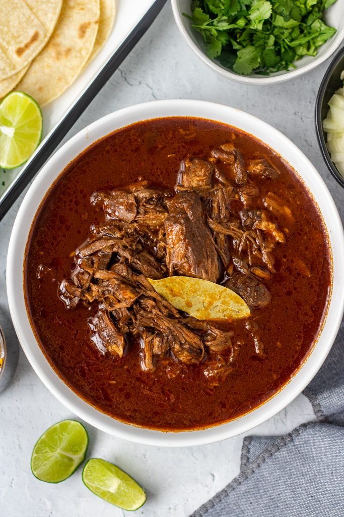 White bowl with birria de res (beef birria) with bay leaf. Lime garnish, bowl of cilantro, bowl of onion, and tortillas in background.