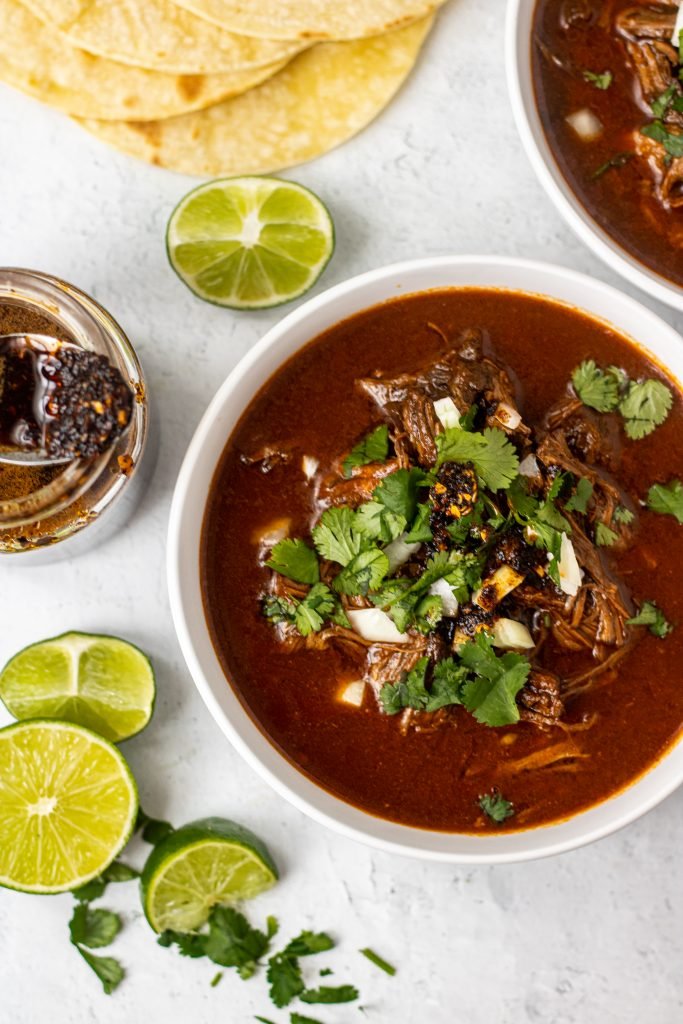 White bowl with birria de res (beef birria) topped with cilantro, onion, and salsa. Lime slices and tortillas on the side.