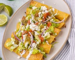 Plate with flautas topped with onion, lettuce, tomato, queso fresco, sour cream, and salsa