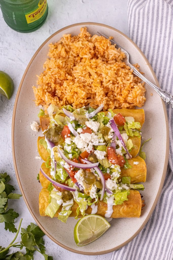 Platter of flautas with side of rice. Flautas topped with lettuce, onion, tomato, queso fresco, sour cream, and salsa.