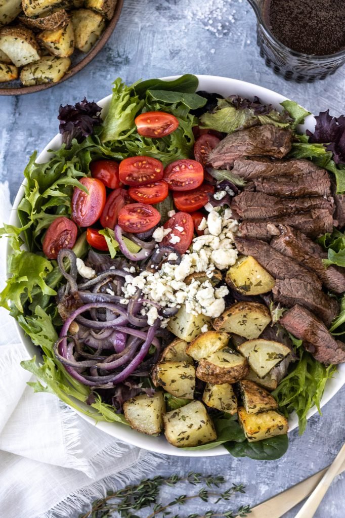 Bowl with steak salad with herbed potatoes, red onions, cherry tomatoes, and gorgonzola. Small container with balsamic vinaigrette dressing